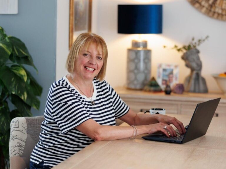 Jayne Porter sitting at a table working at a laptop, turning to smile at the camera