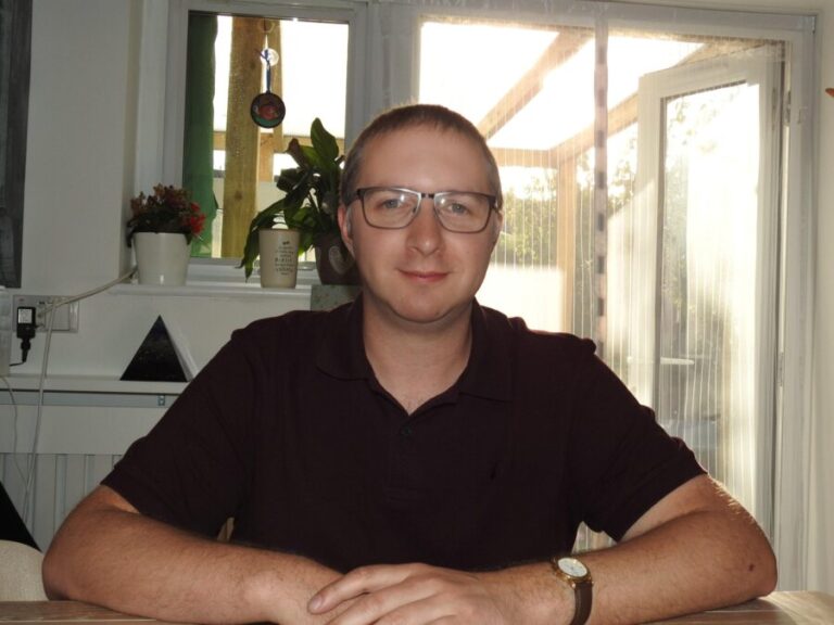 Lloyd Allin sitting at a table in a dark polo shirt and glasses, smiling at the camera